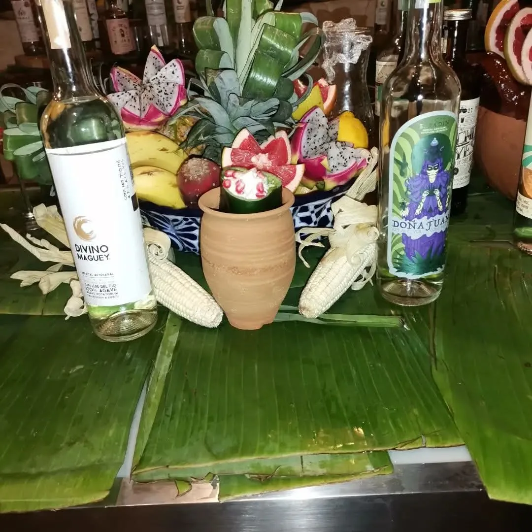 Cóctel en recipiente de barro entre botellas, piñas, pitaya, maíz y hojas verdes.