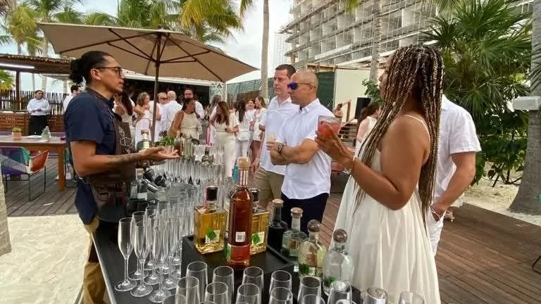 Una persona con mandil conversa con invitados junto a una mesa con copas y botellas al aire libre.