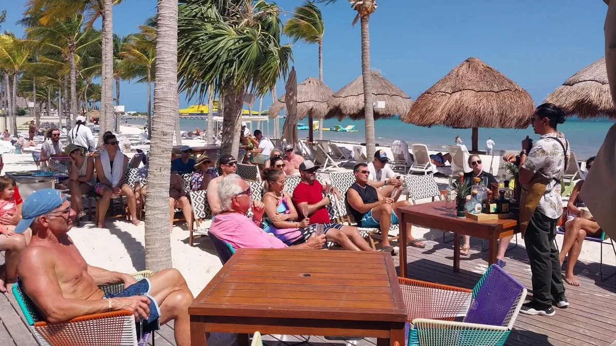 Grupo reunido para una presentación de bebidas junto a una playa con palmeras y sombrillas de palma.