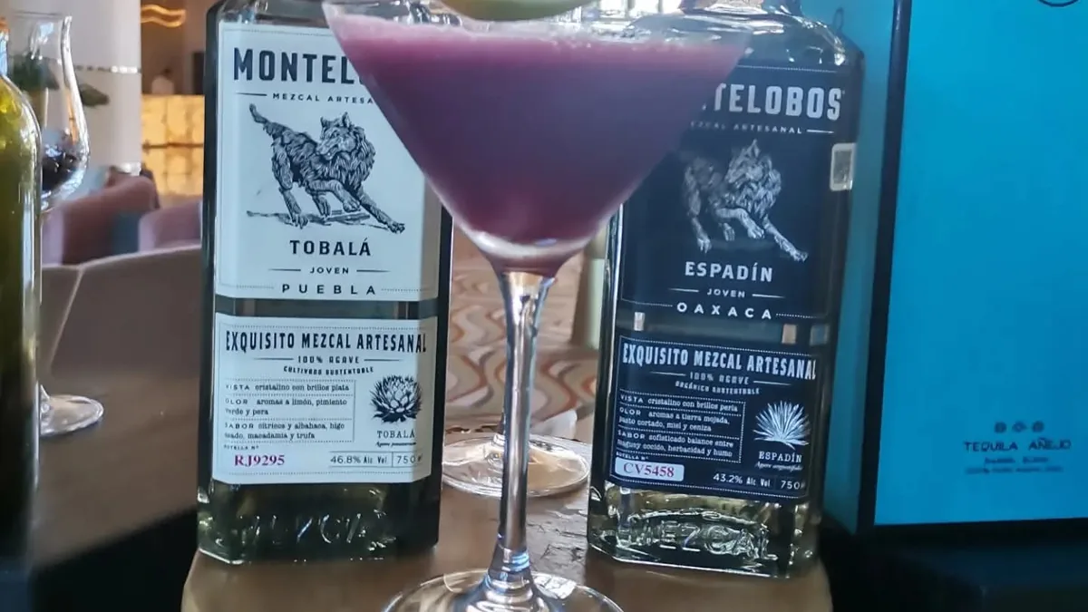Cóctel morado en copa con una hoja verde, junto a botellas de mezcal.
