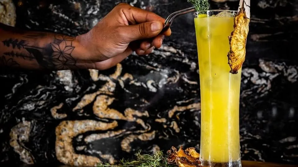 Una mano con tatuajes decora un cóctel amarillo en vaso alto con una pieza de piña dorada.